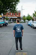 Load image into Gallery viewer, Man standing in city with blue organic cotton tshirt with covid squid design by elk draws